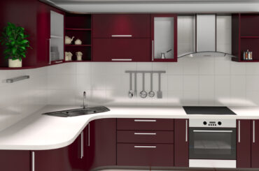 A modern kitchen setup with maroon cabinets, white countertops, a built-in oven, induction cooktop, modular sink, chimney, and stainless steel appliances.